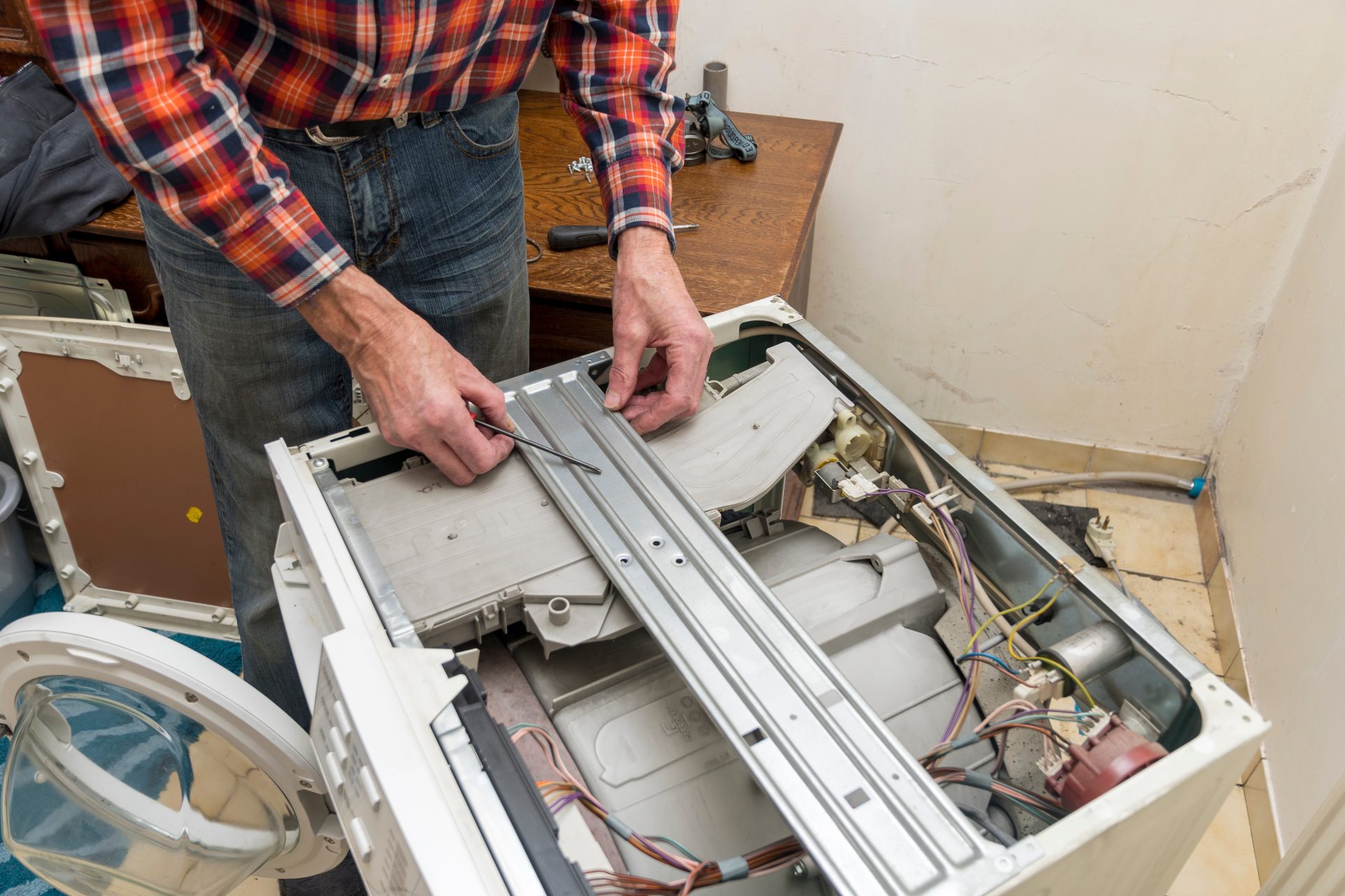 A man repairs a broken washing machine, removes the top cover with a screwdriver